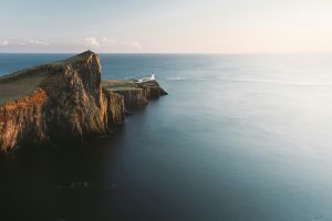 A rocky peninsula jutting out of a calm blue sea with a lighthouse at the end of it.