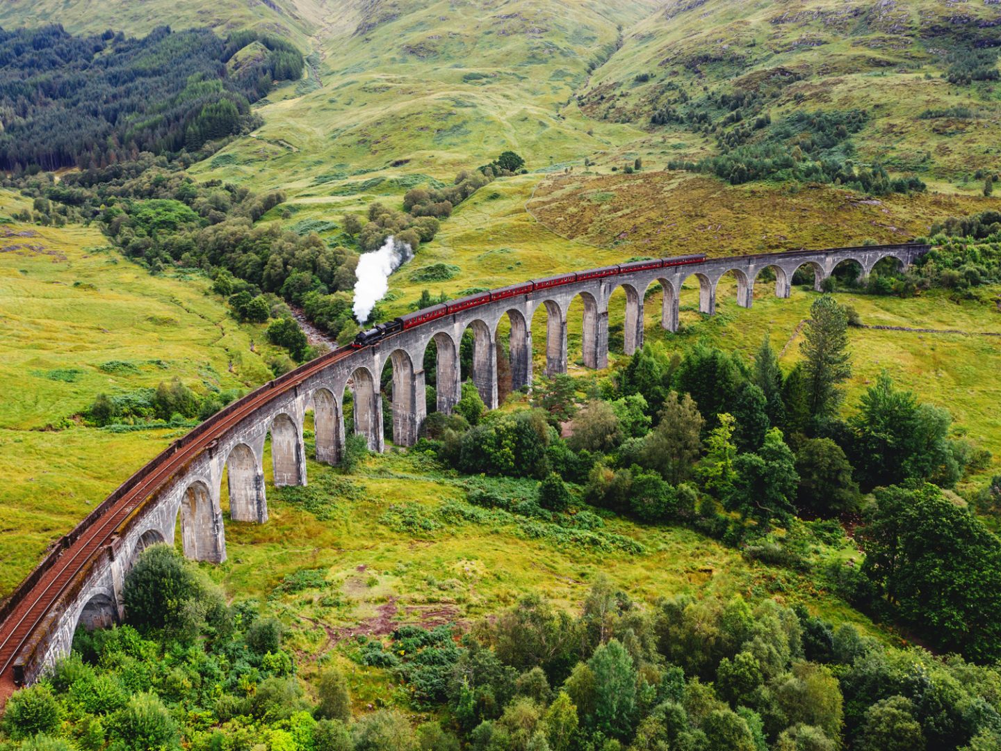 A scenic view of the Jacobite Steam Train crossing the iconic Glenfinnan Viaduct in the Scottish Highlands, surrounded by lush green hills and dense forests.