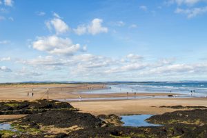 People walking along a wide, sandy beach during daytime with distant hills under a cloudy sky — a peaceful coastal scene ideal for Edinburgh day trips.