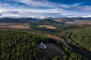 A scenic aerial view of Cairngorms National Park in Scotland, featuring a pyramid-shaped stone structure nestled among dense evergreen forests, with rolling hills and snow-capped mountains in the background under a partly cloudy sky. Ideal for Edinburgh day trips