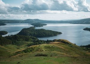Grassy hilltop overlooking a serene lake with rolling hills and scattered trees in the background under a soft, overcast sky in the Scottish Highlands.