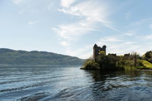 View of Urquhart Castle ruins on a grassy promontory overlooking the calm waters of Loch Ness, with forested hills and a blue sky with wispy clouds in the background.