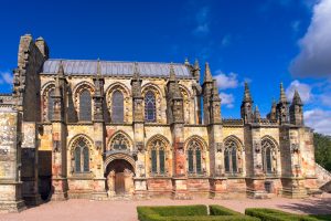 Rosslyn Chapel, a richly decorated 15th-century Gothic church in Midlothian, Scotland, shown on a bright sunny day with intricate stone carvings, arched stained glass windows, and a backdrop of deep blue sky.