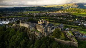 Aerial view of Stirling Castle in Scotland, perched dramatically atop a rocky hill surrounded by lush green trees, with the town of Stirling and rolling hills in the background under a partly cloudy sky.