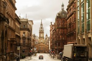 A city street in Glasgow, Scotland, featuring historic sandstone and red-brick buildings lining both sides. In the center background stands the Tolbooth Steeple clock tower, a notable landmark with a pointed spire. The sky is overcast, and cars and pedestrians are visible near the traffic lights.