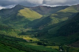 A lush green valley surrounded by rolling hills and dramatic mountain peaks under a partly cloudy sky in the Scottish Highlands, with patches of sunlight casting shadows across the landscape and a winding river visible in the lower right.