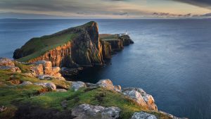 A curved green peninsula with steep cliffsides juts out into a deep blue ocean. At the edge is a white and orange lighthouse, Neist Point, which is one of the best things to see on a 10-Day Scotland itinerary.