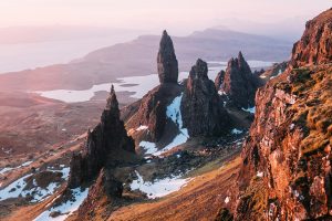 An orange and pink-hued sunset casts rays over four jagged rock pinnacles, the Old Man of Storr, shooting above a hillside in the foreground. One of the must-see places on a 10-day Scotland itinerary.