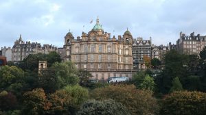 A grand, classical building with a large central dome is pictured in Edinburgh, Scotland. The ornate stone facade rises above a foreground of lush green and autumn-colored trees, with a street and a bus visible at the base.