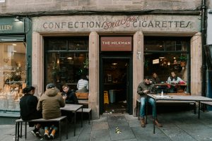 A small coffee shop named "The Milkman" with an old-fashioned sign above the door that reads "CONFECTION SNACKS CIGARETTES." There are people sitting at tables on the cobblestone patio outside and visible through the front windows.
