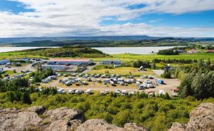 An aerial view of tents, campers, and campervans lining a parking lot surrounded by nature. 