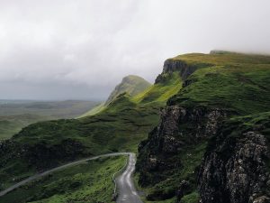A paved road weaves through a moody, verdant landscape with hills.