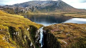 A dramatic aerial shot of the Wailing Widow Falls in Scotland, showing the waterfall plunging through a narrow gorge in the foreground. Above the falls, the water feeds from a tranquil loch reflecting the surrounding rugged, moss-covered mountains. The landscape is dominated by golden-brown and green moorland.