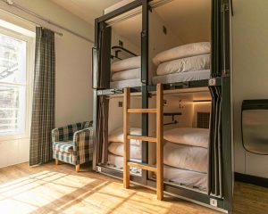 A modern hostel dorm room with four enclosed bunk bed pods featuring individual curtains, lights, and charging outlets. A wooden ladder provides access to the upper bunks. Sunlight streams through a nearby window with plaid curtains, illuminating a plaid armchair. The floor is light wood.