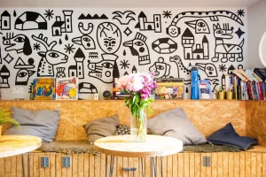 Bright hostel common area in Edinburgh with a quirky black and white mural. Wooden bench with pillows and tables. Board games and flowers add a homey touch.
