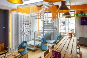 The modern common area of Kick Ass Grassmarket, one of the best hostels in Edinburgh. Features include a long wooden table, spiral staircase, and corrugated metal walls with a large graphic.