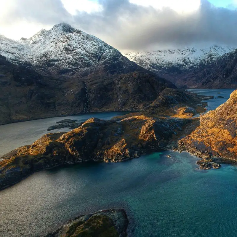 An awe-inspiring winter scene at Loch Coruisk on the Isle of Skye, where snow-capped mountains meet the turquoise edge of the loch. With dramatic light casting golden hues on the rocky terrain, this remote location is a hidden gem worth adding to your Isle of Skye itinerary for unforgettable views and serene solitude.
