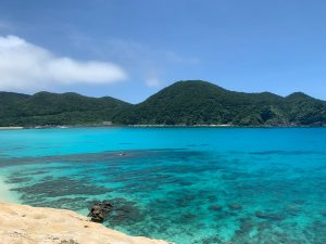 The turquoise waters of Aharen Beach on Tokashiki Island meet against white sand and verdant green mountains in the distance.