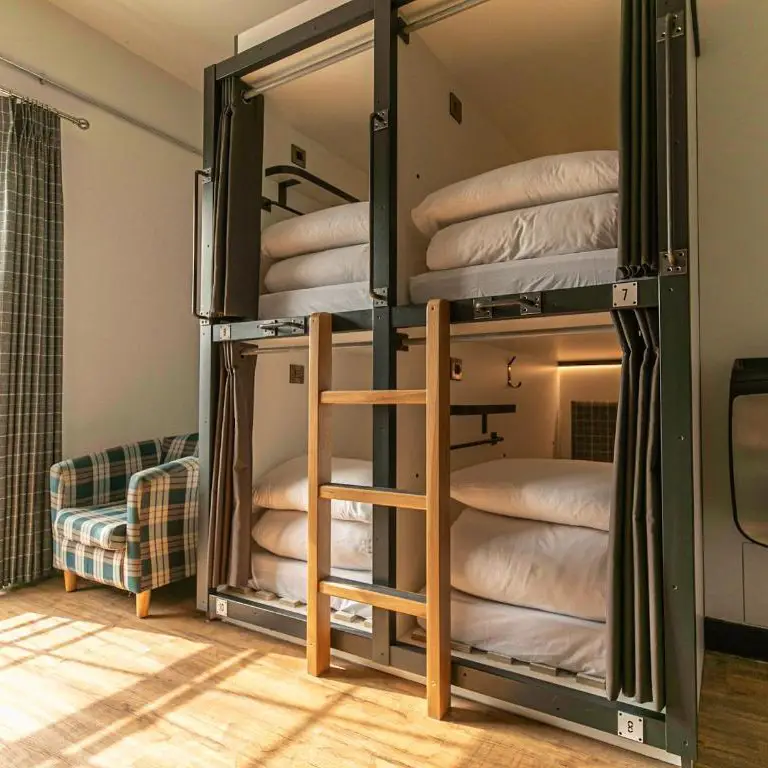 A modern hostel dorm room with four enclosed bunk bed pods featuring individual curtains, lights, and charging outlets. A wooden ladder provides access to the upper bunks. Sunlight streams through a nearby window with plaid curtains, illuminating a plaid armchair. The floor is light wood.