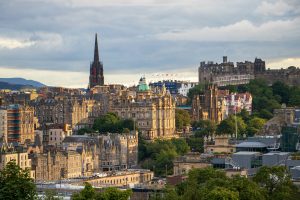 A panoramic view of Edinburgh, Scotland, under a partly cloudy sky. The historic city stretches out with its many stone buildings, a prominent dark spire, and the iconic Edinburgh Castle visible on a hill to the right. This view captures the city's charm, perfect for travelers exploring Scotland on a budget.