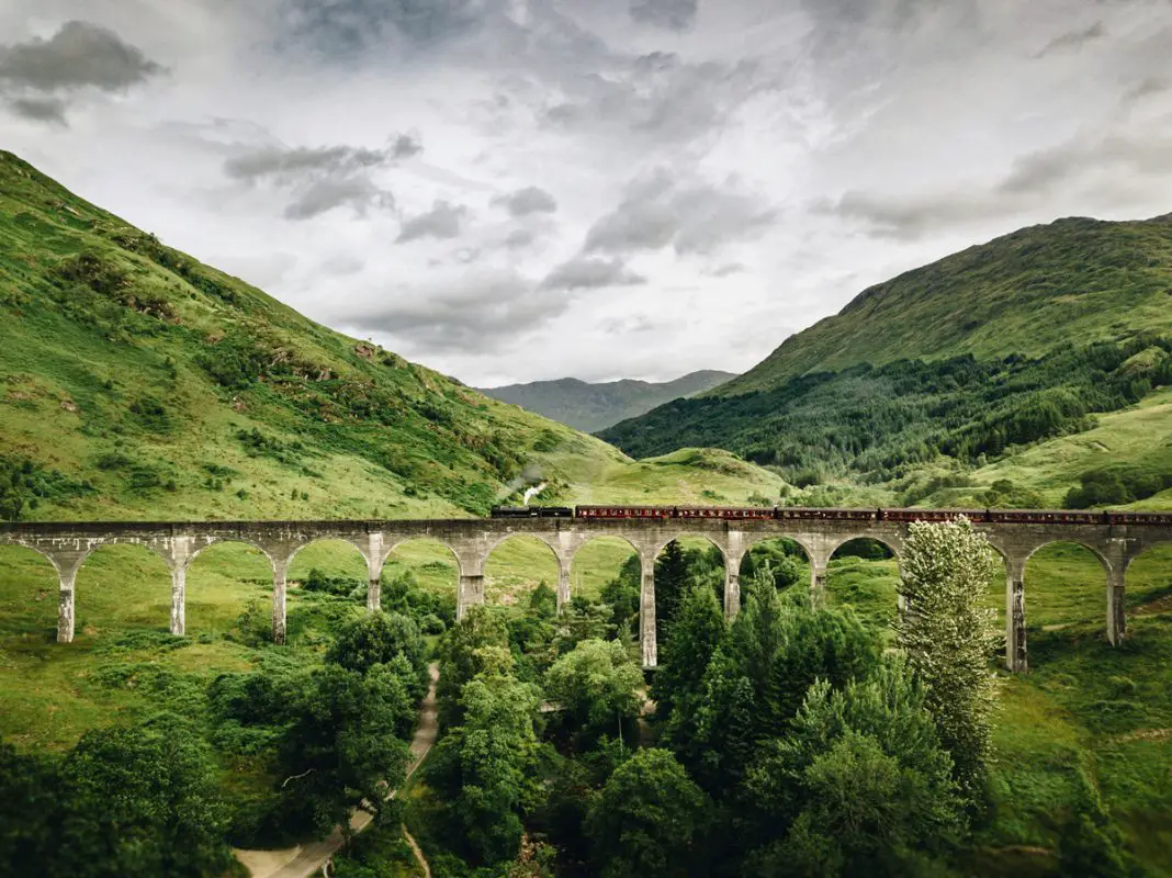 A vintage-style steam train with maroon carriages crossing the Glenfinnan Viaduct in the Scottish Highlands. The viaduct is a long, curved stone bridge supported by multiple arches, set against a dramatic backdrop of steep, green, tree-covered mountains under a cloudy sky.