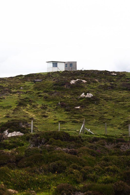 Small white bothy on a grassy hilltop above a fenced slope of heather.
