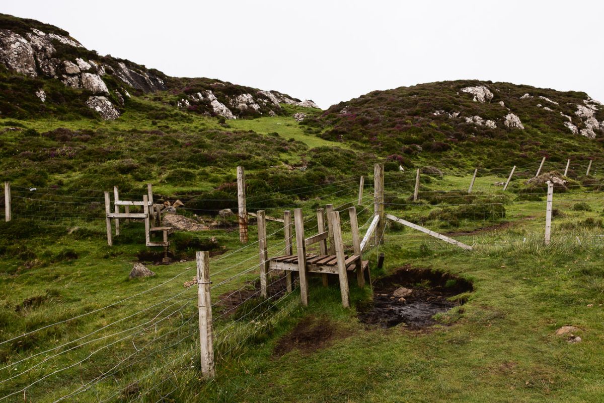 Wooden stile over a wire fence with rocky heather-covered hills behind.
