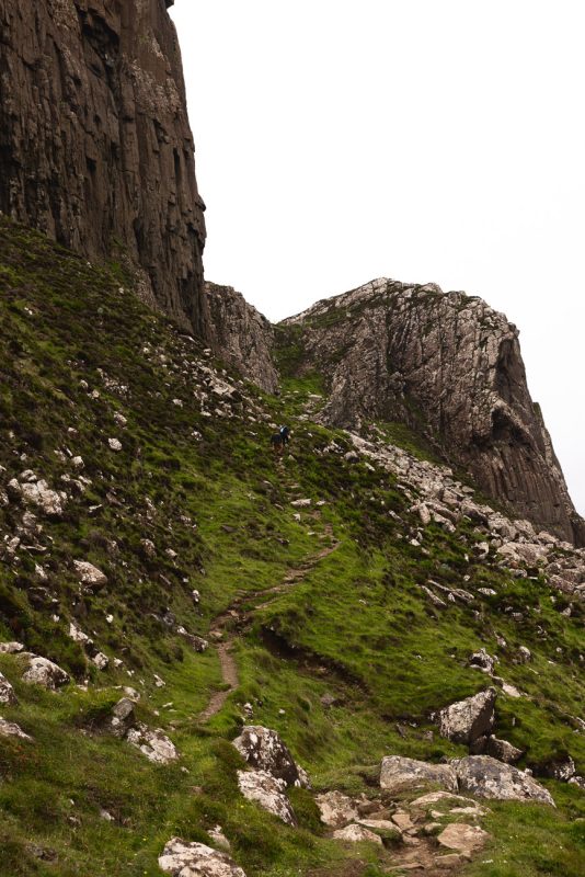 Two hikers walking a narrow path up a steep grassy slope beneath dark cliffs.
