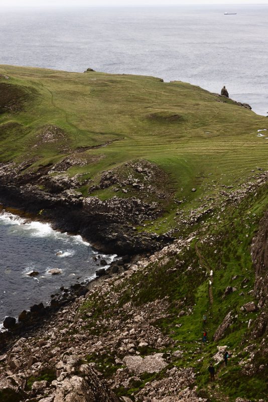 Tiny hikers on a narrow path beside a rocky cove, with a grassy headland and open sea beyond.