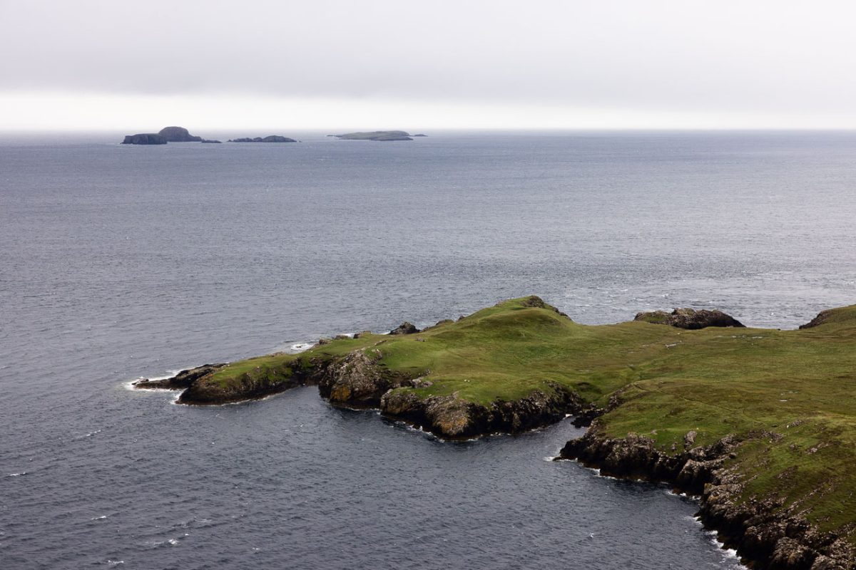 Green headland jutting into dark water with small islands on the horizon.