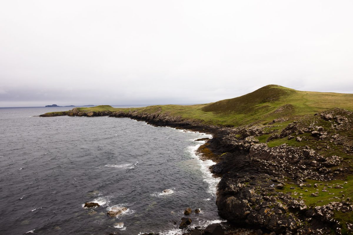 Waves breaking along a dark rocky shoreline below a green headland.