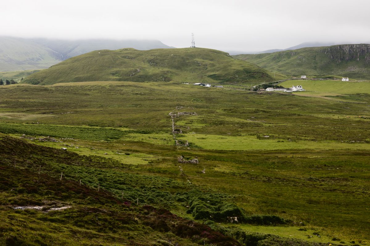 Green moorland with a ruined stone wall and white farmhouses below a hill topped by a radio mast.